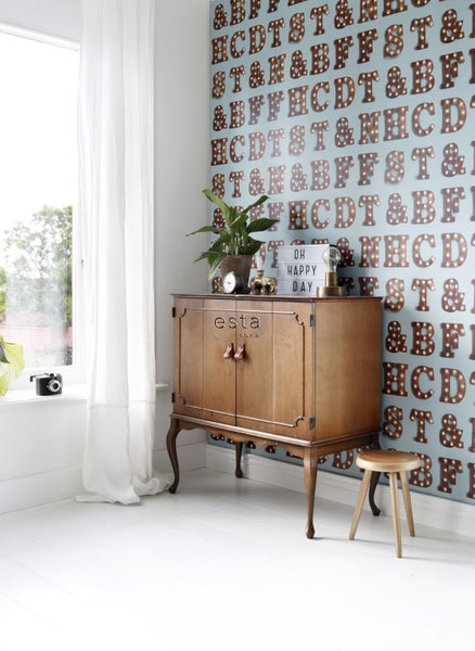 Wohnzimmer mit Holzkommode, Hocker und Wandtapete mit Buchstabenmuster