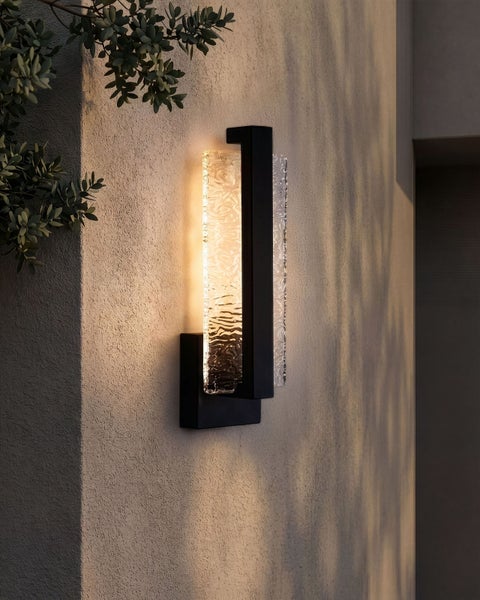 Moderne Außenwandleuchte mit schwarzem Metallrahmen und strukturiertem Glas, montiert an einer Fassade mit warmem Lichtschein.