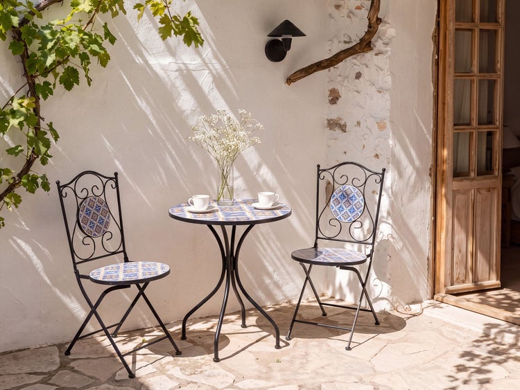 Bistrotisch Set mit zwei Stühlen und einem runden Tisch aus Metall mit mediterranen Kacheleinlagen auf einer Terrasse vor einer weißen Mauer.