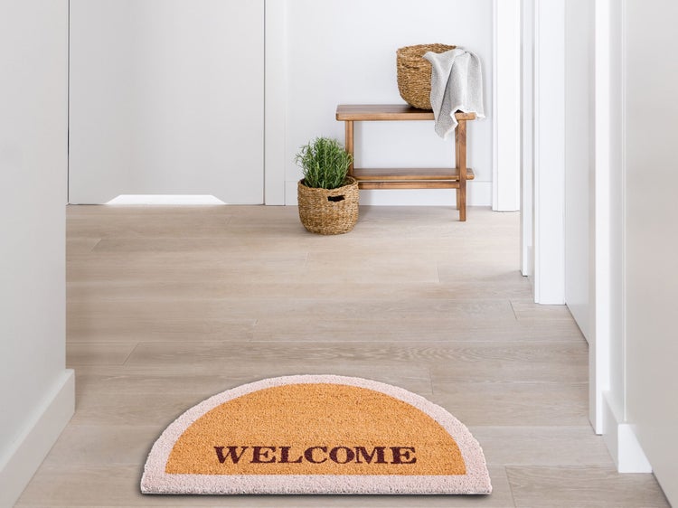 Halbmondförmige Fußmatte mit Welcome-Schriftzug in einem hellen Flur mit Holzbank und geflochtenen Körben.