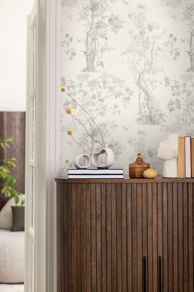 Vliestapete mit floralem Baum- und Vogelmuster im Landhausstil vor einer Kommode aus geriffeltem Holz mit Dekorationsartikeln.
