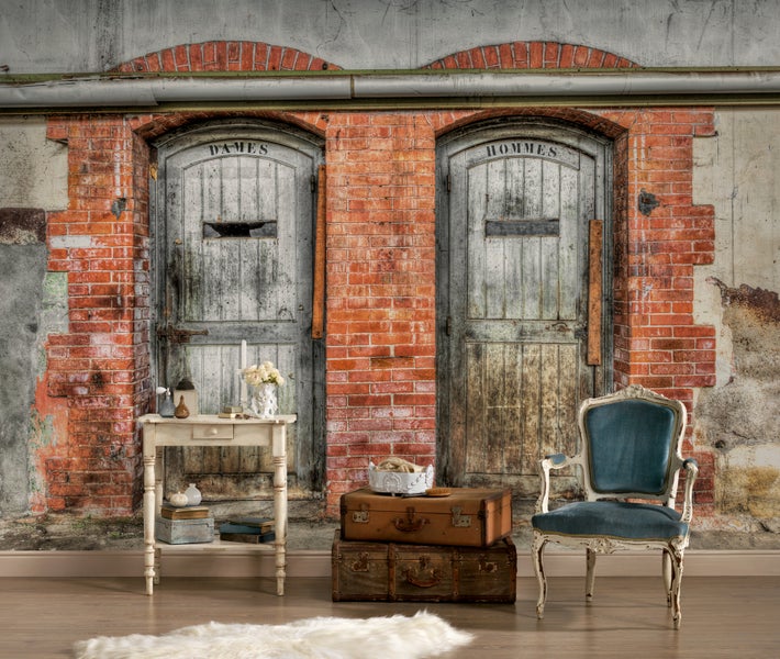 Fototapete mit Backsteinwand und zwei rustikalen Holztüren im Industriedesign, davor stehen ein Sessel, Koffer und ein kleiner Beistelltisch.