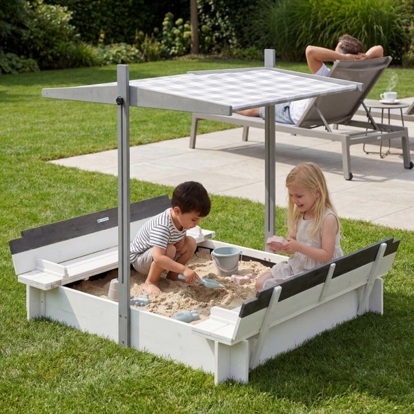 Moderner weißer Holz-Sandkasten im Garten mit zwei spielenden Kindern, verstellbarem Karodach und integrierten Sitzbänken.