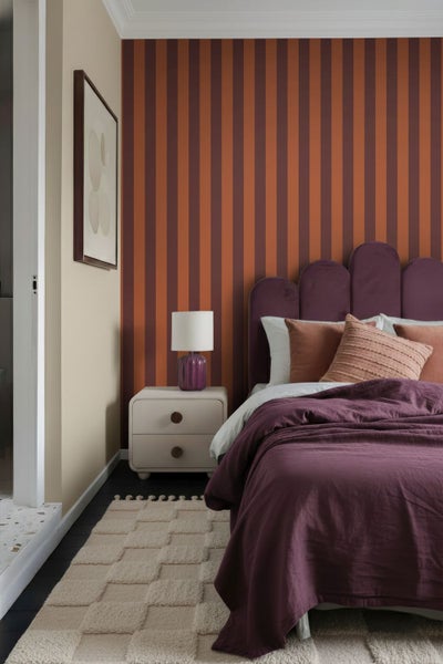 Modernes Schlafzimmer mit vertikal gestreifter Tapete in Terrakotta und Dunkelrot, lila Polsterbett mit bogenförmigem Kopfteil und hellem Nachttisch.