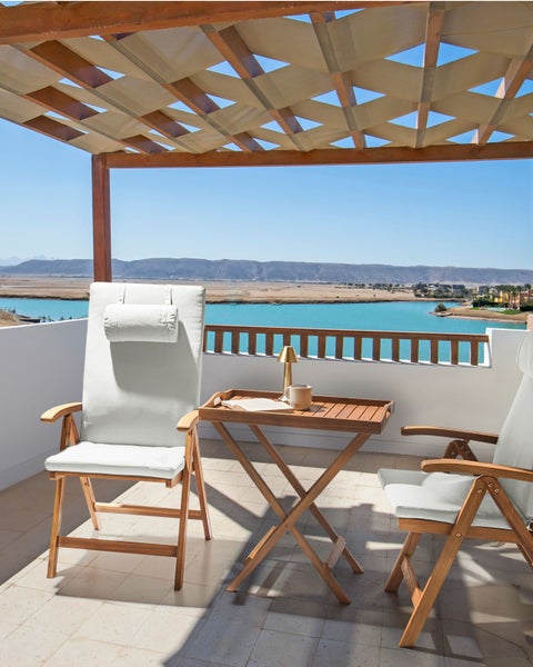 Hölzerne Gartenmöbel auf einer hellen Terrasse unter einer Pergola, zwei Klappsessel mit weißen Polstern und ein Klapptisch vor Meereskulisse.