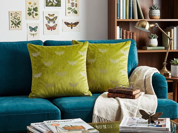 Zwei quadratische Kissen mit Schmetterlingsmuster auf einem Sofa in einem gemütlich eingerichteten Wohnzimmer mit Regalen und Dekoration.