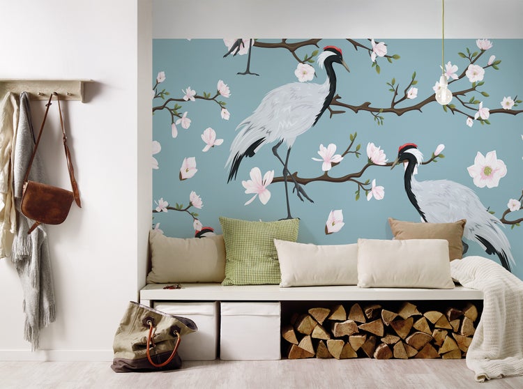 Fototapete mit Kranichen und Blütenzweigen über einer Sitzbank mit Kissen und Kaminholz in einem hellen Zimmer.