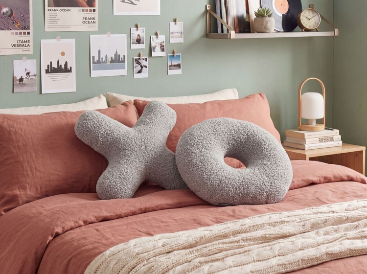 Modernes, helles Schlafzimmer mit lachsfarbener Bettwäsche, grauen Teddy-Dekokissen in X- und O-Form, Strickdecke, Wandregal und Nachttisch.