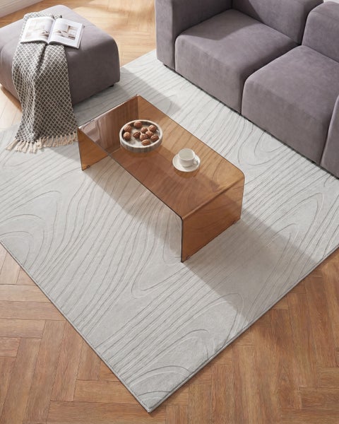 Moderner Teppich mit Wellenmuster in Hellgrau, liegend in einem hellen Wohnzimmer unter einem Couchtisch aus Glas neben einem Sofa.