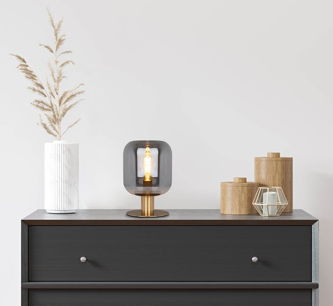 Dekorative Szene mit Tischleuchte, Vase, Trockenblumen und Aufbewahrungsdosen auf einer Kommode.