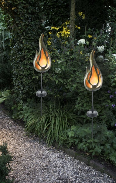 Zwei Gartenleuchten in Flammenform entlang eines Kieswegs inmitten von Grünpflanzen.