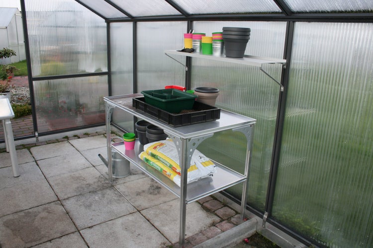 Aluminium-Pflanztisch mit zwei Ablagen und passendem Wandregal in einem Gewächshaus mit Blumentöpfen und Blumenerde.
