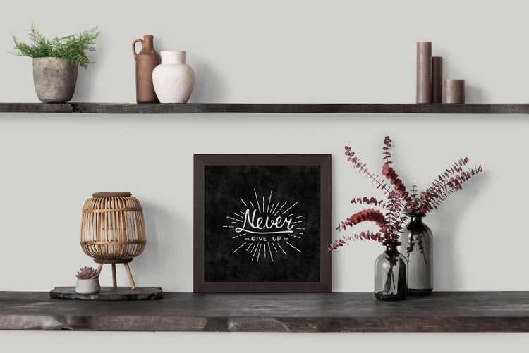 Quadratisches Wandbild Never GIVE UP in dunklem Holzrahmen auf einem Holzregal mit Dekoration wie Pflanzen, Vasen und einer hölzernen Leuchte.