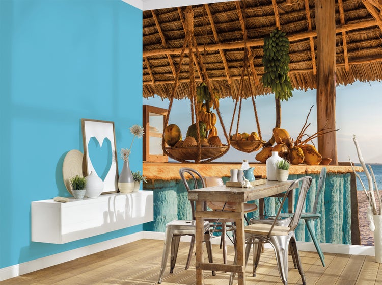 Fototapete mit tropischer Strandbar, Reetdach und Früchten in einem Esszimmer mit Holztisch und blauer Wand.