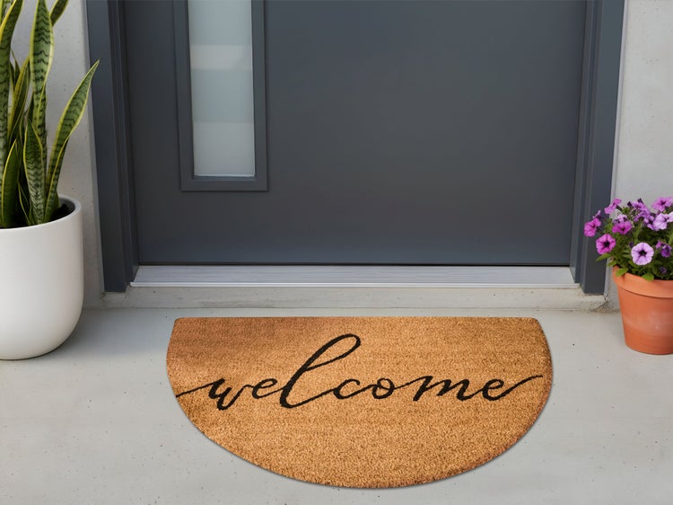 Halbmondförmige Kokos-Fußmatte mit Welcome-Schriftzug vor einer modernen grauen Haustür, flankiert von Topfpflanzen auf Betonboden.