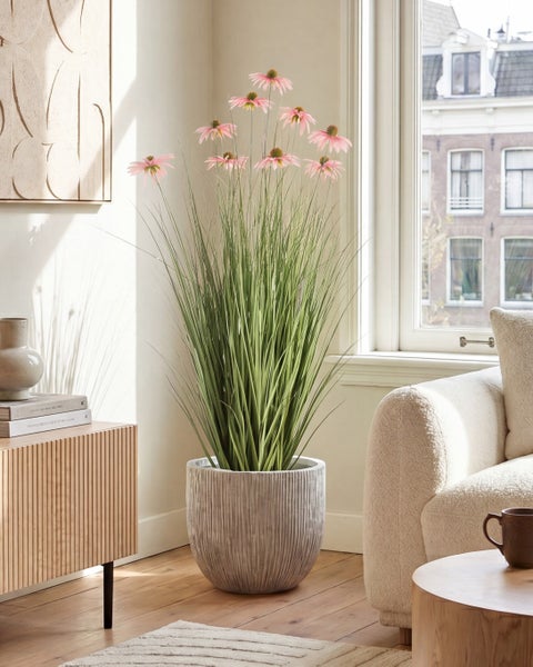 Künstliches Gras mit rosa Blüten im grauen Topf, arrangiert in einem hellen, modernen Wohnzimmer neben Sofa und Holzsideboard.