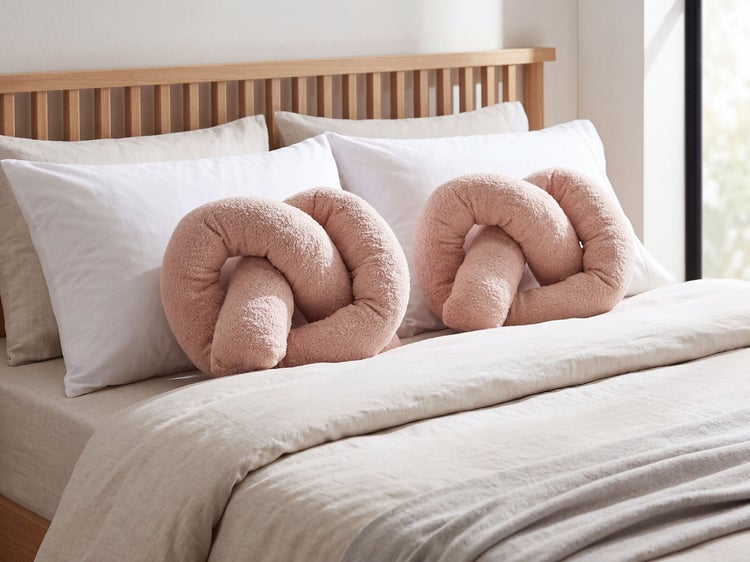 Zwei dekorative Knoten-Kissen aus strukturiertem Stoff liegen auf einem Bett mit Holz-Kopfteil und weißer Bettwäsche.
