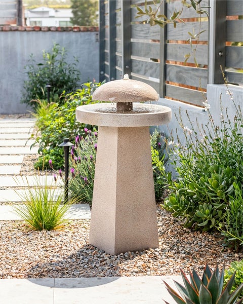 Moderner Gartenbrunnen in Steinoptik mit pilzförmigem Aufsatz, platziert in einem Kiesbeet umgeben von verschiedenen Gartenpflanzen.