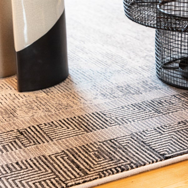 Nahaufnahme eines Teppichs mit geometrischem Muster in Grau und Schwarz auf Holzboden neben einer Vase und einem Drahtkorb.