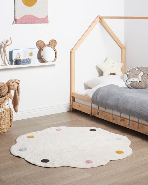Helles Kinderzimmer mit weißem, gewelltem Teppich mit bunten Punkten, einem Hausbett aus Holz und dekorativen Accessoires.