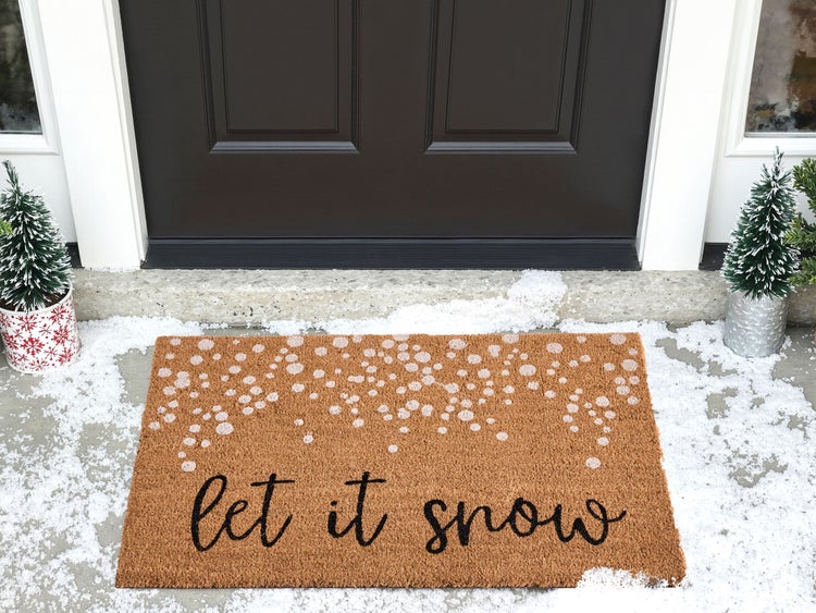 Fußmatte mit der Aufschrift "Let it snow" vor einer Haustür mit zwei kleinen Weihnachtsbäumen