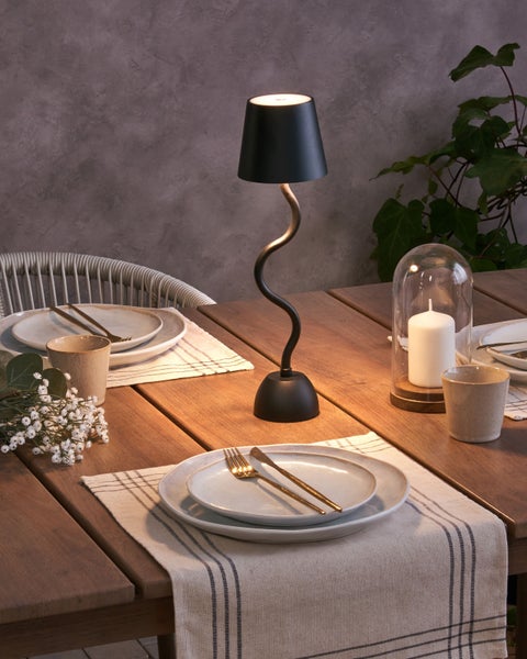 Moderne LED-Tischleuchte mit geschwungenem Fuß auf einem gedeckten Holztisch mit Geschirr, Besteck und Kerze in stimmungsvollem Ambiente.