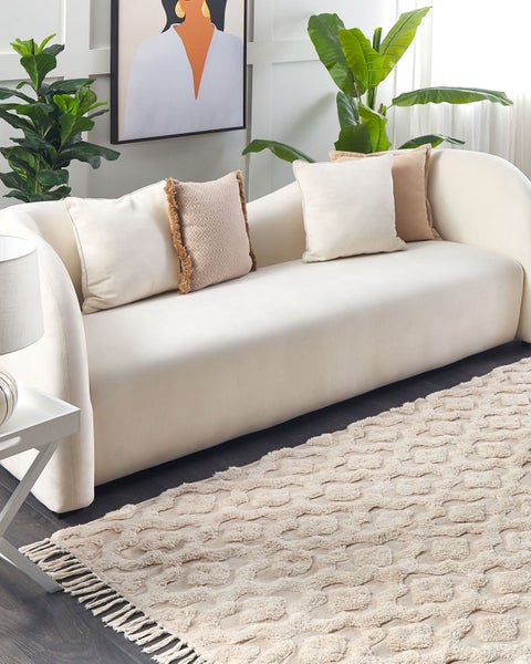 Modernes Wohnzimmer mit cremefarbenem geschwungenem Sofa, Zierkissen, gemustertem Teppich mit Fransen und grünen Zimmerpflanzen.