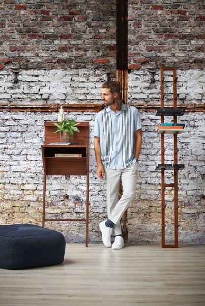 Mann steht neben einem Holzsekretär und einem Holzregal in einem Zimmer mit Ziegelwand