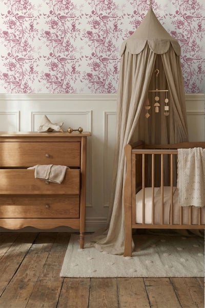 Klassisches Kinderzimmer mit Holzbett, Kommode und Tapete mit Vogelmuster. Ein Betthimmel aus Stoff und helle Textilien schaffen ein helles Ambiente.