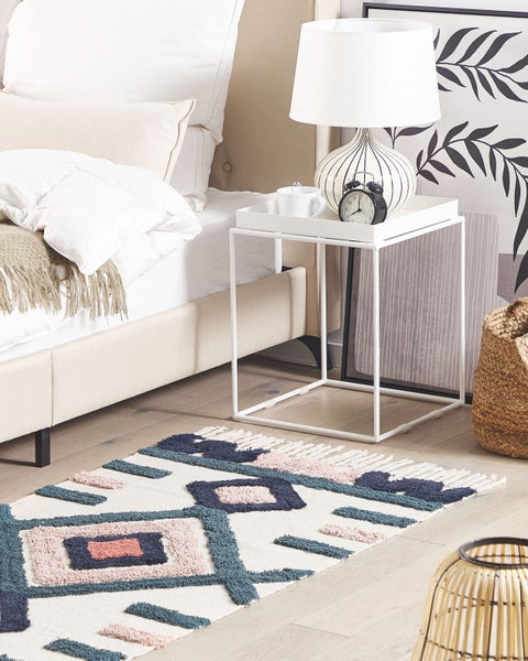 Hell gestaltetes Schlafzimmer mit weißem Metallbeistelltisch neben dem Bett, gemustertem Teppich, Nachttischlampe und Dekoration.