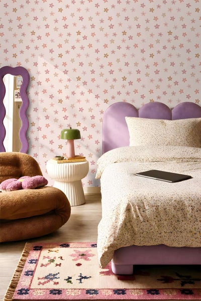 Tapete mit Blumenmuster in einem eingerichteten Schlafzimmer mit violettem Bett, gewelltem Spiegel und einem gemusterten Teppich auf dem Boden.