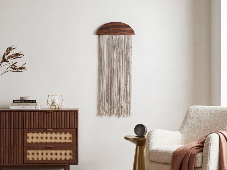 Hell gestalteter, minimalistischer Wohnraum mit einem Makramee Wandbehang aus Holz und Textil, einem Sideboard mit Rattan und einem Sessel.