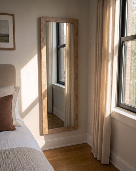 Ganzkörperspiegel mit hellem Holzrahmen an einer hellen Wand im Schlafzimmer neben einem Fenster mit Vorhängen.