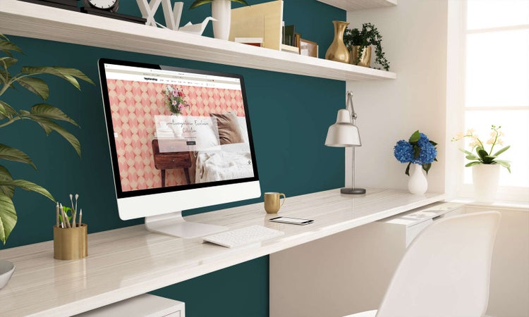 Modernes Arbeitszimmer mit petrolfarbener Wand, hellem Schreibtisch, Monitor mit Tapetenmuster, Schreibtischlampe und Wandregalen.