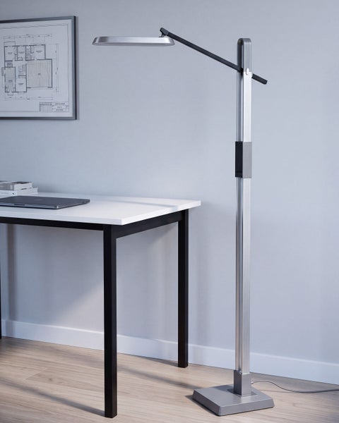 Moderne Stehleuchte aus Metall mit flachem Leuchtenkopf neben einem Schreibtisch in einem schlicht eingerichteten Arbeitszimmer.
