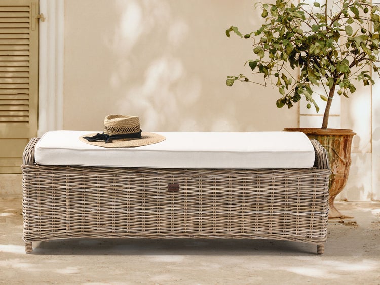 Geflochtene Gartenbank aus Rattan mit weißem Sitzkissen und Strohhut neben einem Pflanztopf auf einer hellen Terrasse.