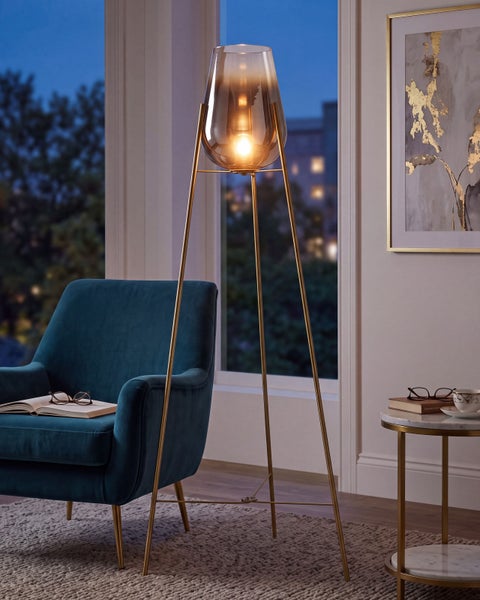 Moderne Stehleuchte mit dreibeinigem Metallgestell und tulpenförmigem Glasschirm in einem gemütlichen Wohnzimmer mit Sessel und Beistelltisch.