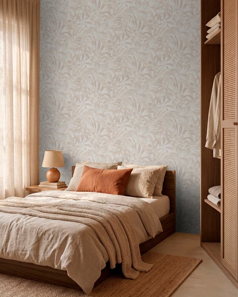 Schlafzimmer mit heller Blättermuster-Tapete an der Wand hinter einem Holzbett mit beigen Textilien und einem rostfarbenen Kissen.