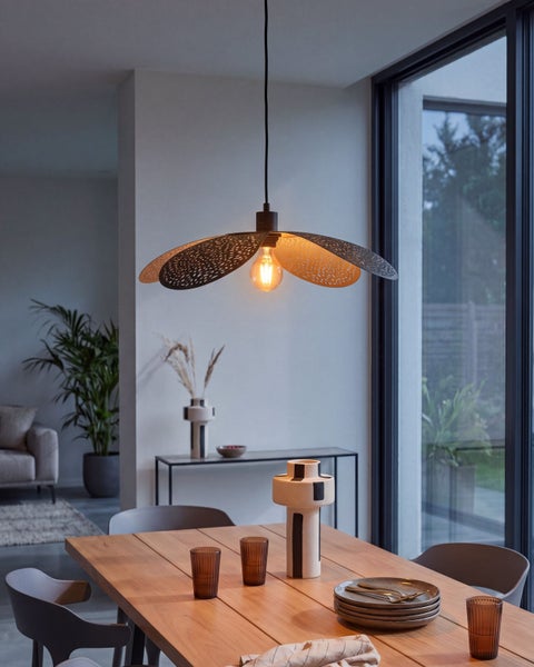 Moderne Pendelleuchte mit perforiertem Metallschirm in Blütenform über einem Holztisch in einem hellen, zeitgemäßen Raum mit großem Fenster.