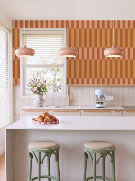 Helle Küche mit geometrischer Tapete in Orange, Kochinsel, grünen Barhockern aus Holz und drei rosa Pendelleuchten über der Arbeitsplatte.