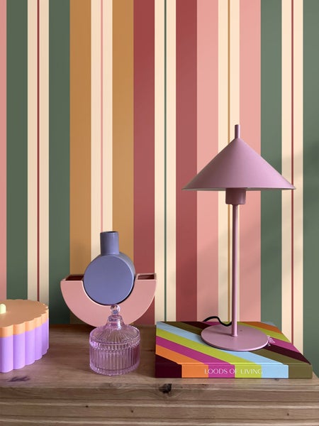 Vertikal gestreifte Tapete in verschiedenen Farben wie Grün, Rosa und Ocker hinter einem Sideboard aus Holz mit dekorativer Tischlampe und Vasen.