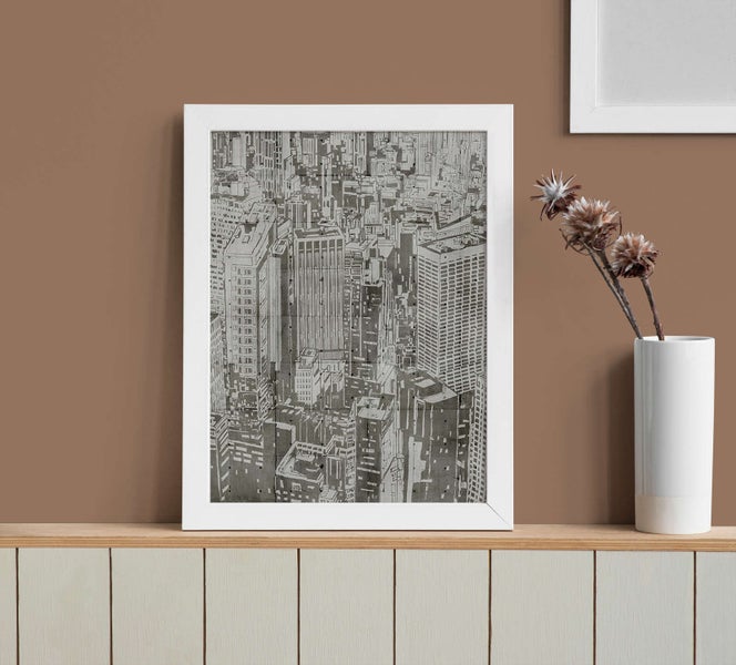 Gerahmtes Wandbild mit einer schwarz-weißen Illustration einer Skyline in einem weißen Rahmen auf einem Regal.