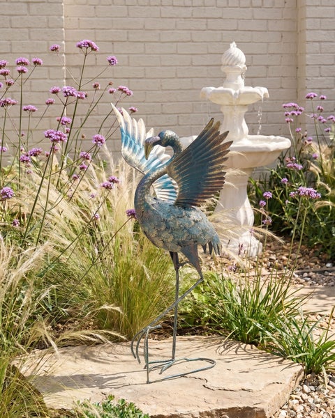 Gartendekoration Kranich aus Metall mit ausgebreiteten Flügeln in einem Garten mit Gräsern, lila Blumen und einem weißen Springbrunnen.