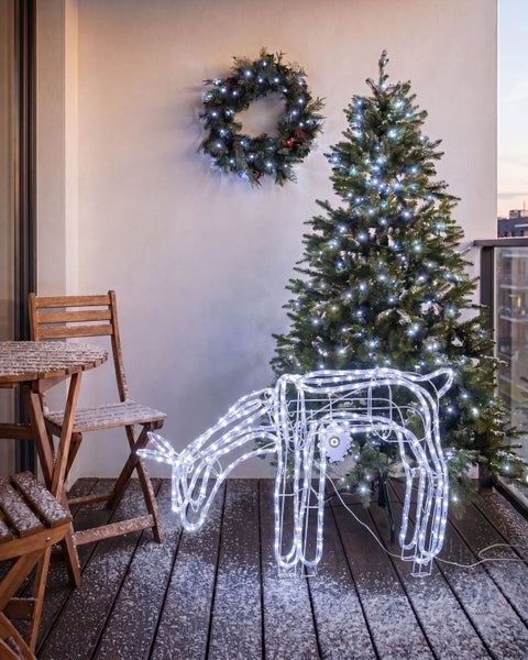 Festlich dekorierter Balkon mit beleuchtetem Weihnachtsbaum, LED-Rentier, Kranz und schneebedeckten Holzmöbeln.