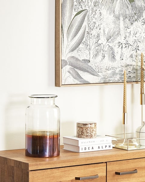 Dekorative Szene mit einer Glasvase, Büchern und Kerzen auf einer Holzkommode unter einem gerahmten Bild.