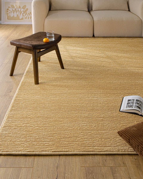 Großer strukturierter Teppich mit horizontalem Rippenmuster in einem hellen Wohnzimmer auf Holzfußboden, neben Sofa und Hocker.