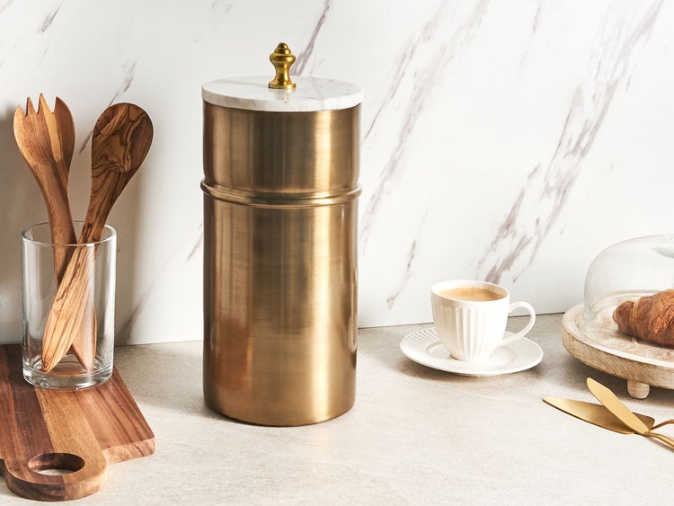 Zylindrische goldfarbene Vorratsdose mit weißem Marmordeckel und goldenem Knauf in einer hellen Küche neben Besteck und Kaffeetasse.