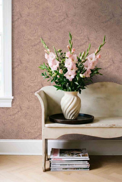 Raumszene mit floral gemusterter Tapete, einer geschwungenen Holzbank mit Blumenvase und einem Zeitschriftenstapel auf Fischgrätparkett.