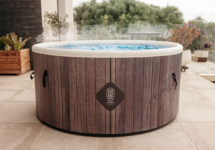 Runder aufblasbarer Whirlpool in dunkler Holzoptik mit weißem Rand und sprudelndem Wasser auf einer Terrasse. COCO SPA Logo.