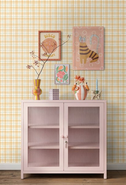 Modernes Wohnzimmer mit einem rosafarbenen Metallschrank mit Glastüren vor einer gelb-weiß karierten Tapete und bunter Dekoration.